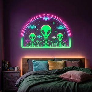 Puede incluir: Un letrero de neón semicircular para pared que presenta tres figuras alienígenas verdes centradas bajo un marco arqueado rosa y azul. El diseño incluye pequeños planetas y estrellas brillantes que rodean a los alienígenas.