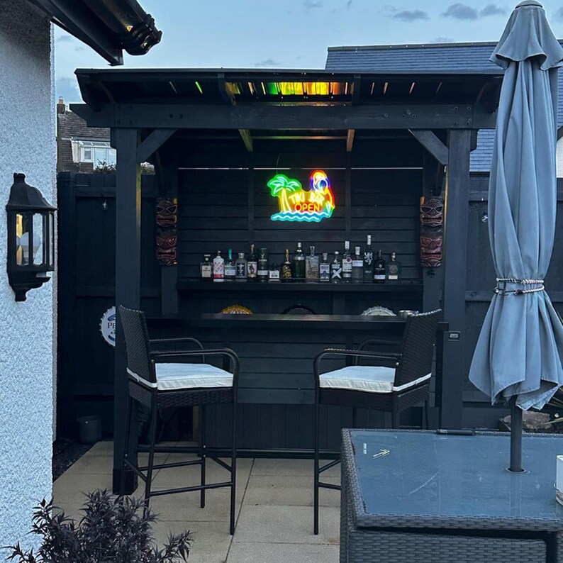 May include: A black wooden outdoor bar structure featuring a neon sign that reads Tiki Bar Open, two wicker bar stools with white cushions, a shelf displaying various glass liquor bottles, and two decorative tiki masks mounted on the side posts.