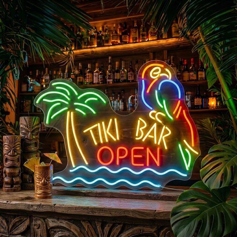 May include: A neon sign featuring a palm tree, a parrot, and the text TIKI BAR OPEN in bright colours. The sign is displayed on a wooden surface next to a brown ceramic tiki mug with a small cocktail umbrella, surrounded by green tropical leaves.
