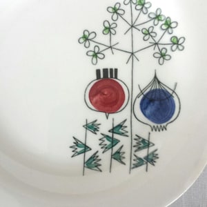 Puede incluir: Un plato de cerámica blanco con un diseño floral estilizado en negro, verde, rojo y azul. El diseño presenta una flor con un tallo y hojas, un círculo rojo con una franja negra y un círculo azul con un contorno negro.