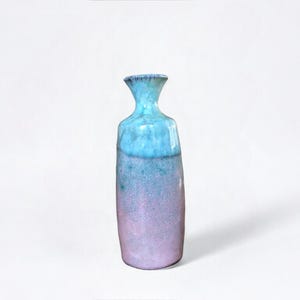 May include: A tall, ceramic vase with a unique shape and a gradient color scheme. The top of the vase is a bright turquoise, transitioning to a lighter blue, and then to a soft lavender at the base. The vase has a textured surface.