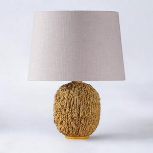 Może przedstawiać: Lampa stołowa z beżowym abażurem z lnu i okrągłą, teksturowaną podstawą wykonaną z naturalnych materiałów. Podstawa jest jasnobrązowa i ma szorstką, organiczną fakturę.