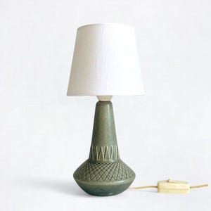 May include: A vintage table lamp with a white lampshade and a green ceramic base. The base has a textured diamond pattern and a decorative band. The lamp includes a cord and a switch. The lamp is approximately 12 inches tall.