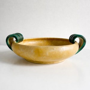 Puede incluir: Un cuenco de cerámica amarilla con dos asas verdes y curvas. El cuenco tiene un diseño ancho y poco profundo y una superficie texturizada. Las asas son de un tono verde más oscuro y añaden un elemento decorativo.