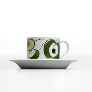 May include: A white ceramic coffee cup with a green and black abstract design. The cup is sitting on a white saucer.