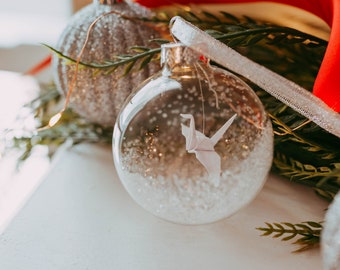 Könnte beinhalten: Eine transparente Glaskugel mit weißem Kunstschnee und einem weißen Origami-Kranich. Der Schmuck hängt an einem dünnen Faden. Weitere Ornamente und Grünzeug befinden sich im Hintergrund.