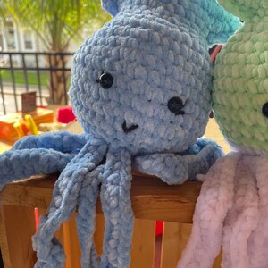 May include: Two crocheted octopus plushies, one light blue and one light green, with black eyes and a smiling mouth. The plushies are sitting on a wooden surface.