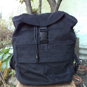 Op de afbeelding: Een zwarte corduroy rugzak met een klepsluiting en een zwarte plastic gesp. De rugzak heeft een voorvak en verstelbare schouderbanden. Hij staat op een kleine houten kruk.