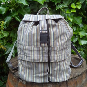 Op de afbeelding: Een gestreepte rugzak met verticale strepen in tinten blauw, bruin en crème. De rugzak heeft een klepsluiting, een zwarte gesp en verstelbare bandjes. Hij staat op een houten vat.