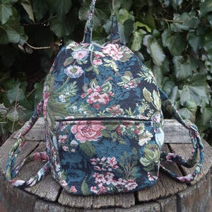 Könnte beinhalten: Ein Rucksack aus dunkelblau-grünem Stoff mit einem gewebten Blumenmuster in Rosa-, Weiß- und Grüntönen. Die Tasche verfügt über einen Tragegriff, zwei verstellbare Schultergurte und eine horizontale Reißverschlusstasche an der Vorderseite.