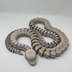 May include: A 3D printed snake sculpture made of layered plastic. The snake is coiled in a relaxed pose with its head raised. The sculpture is painted in shades of brown, white, and purple.