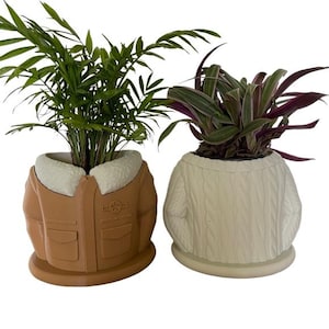 Peut inclure: Deux pots en céramique en forme de veste et de pull. Le pot en forme de veste est brun avec un col en fausse fourrure blanche et le pot en forme de pull est blanc avec un motif en tricot torsadé. Les deux pots ont des plantes qui poussent à l'intérieur.