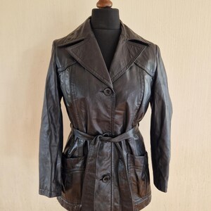 May include: A dark brown leather jacket with a belted waist and two front pockets. The jacket has a notched collar and button closures. The jacket is displayed on a mannequin.