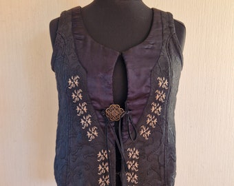 Op de afbeelding: Een zwart, mouwloos vest met een satijnen voering en een decoratieve knoopsluiting. Het vest is geborduurd met een bloemenpatroon in goudgaren.