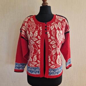 Puede incluir: Cárdigan rojo con estampado floral blanco. El suéter tiene cuello redondo, mangas largas y un diseño geométrico azul y blanco en los puños. Se exhibe en un maniquí negro, con un ribete negro.