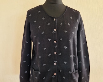 May include: Dark navy cardigan with a delicate floral print. The button-up sweater features two front pockets and is displayed on a mannequin. The cardigan is a dark colour with a subtle, light-coloured floral design.