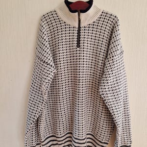 May include: A cream and black patterned sweater with a high collar and a quarter-zip. The sweater features a grid design and black stripes at the hem. The collar is lined with a dark red fabric.