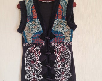 Op de afbeelding: Een zwart, mouwloos vest met een kleurrijk geborduurd ontwerp. Het vest heeft een V-hals en een knoopsluiting. De borduursels bevatten bloemen- en paisleypatronen in tinten blauw, rood, oranje en wit.