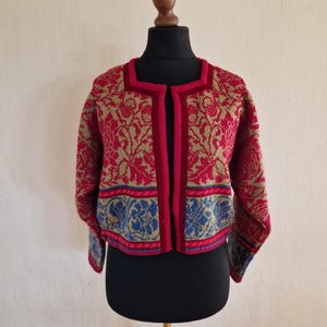 Pode incluir: Cardigã vermelho e dourado estampado, exibido em um manequim. O suéter aberto apresenta um design floral com detalhes azuis na bainha. A borda e o forro interno são de cor vermelha lisa. A peça é um estilo cropped.