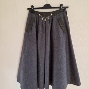 Puede incluir: Falda de lana gris con silueta en forma de A. La falda presenta una cintura y detalles de bolsillo en verde oscuro. Una cadena plateada con dijes circulares cuelga del frente. La falda está colgada en una percha negra.