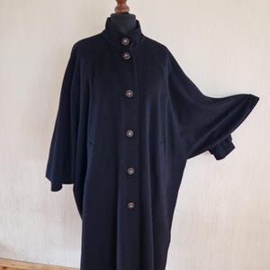 May include: A black, button-down coat with a high collar and wide sleeves. The coat is displayed on a mannequin and features large, round buttons down the front. The coat's design is simple and elegant.