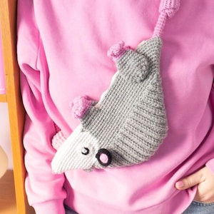 Puede incluir: Un bolso bandolera con forma de zarigüeya de ganchillo gris y blanco con detalles en rosa. El bolso tiene detalles de ojos negros y una correa larga. El bolso se lleva sobre una sudadera rosa y unos vaqueros azules.