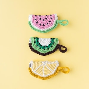 May include: Three crocheted fruit-shaped coin purses: a watermelon with pink, green, and black details, a kiwi with green, brown, and white, and a lemon with yellow and white. Each purse has a loop handle and a zipper.