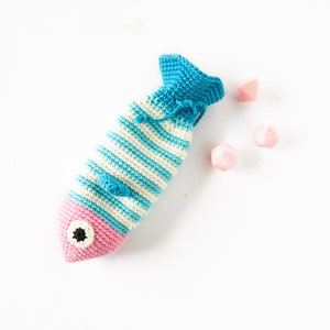 May include: A crocheted fish-shaped bag with blue and white stripes and a pink face. The bag is filled with pink dice.