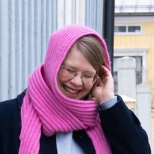 May include: A woman wearing a pink knitted scarf and a blue jacket. She is smiling and looking down.