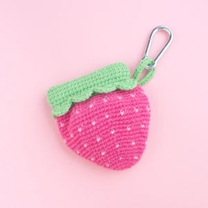 May include: A handmade crochet strawberry-shaped pouch in bright pink with white seed details and a green leafy top. It has a silver carabiner clip attached for easy carrying. The pouch is set against a soft pink background.