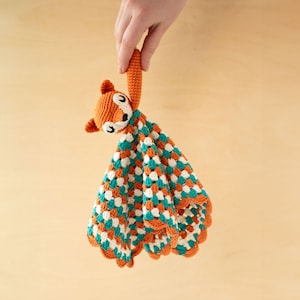 May include: A crocheted fox security blanket with orange and teal colors. The blanket is made with a soft, textured yarn and has a cute fox face with black eyes and a pink nose.