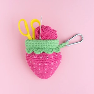Op de afbeelding: Een gehaakte aardbeivormige etui met een groene rand en een zilveren karabijnhaak. De etui bevat een gele schaar, een haaknaald en een bol roze garen. De aardbei is roze met witte stippen.