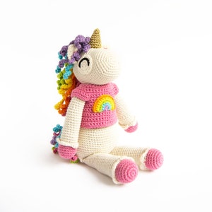 以下が含まれることがあります： 虹色のたてがみと虹のデザインのピンクのシャツを着た、かぎ針編みで作ったユニコーンのぬいぐるみ。ユニコーンは白い背景に座っています。