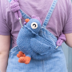 Puede incluir: Bolso azul tejido a ganchillo con forma de pollo, con patas naranjas y cresta morada. El bolso tiene un ojo blanco y negro y está sujeto a una correa morada. El bolso se lleva sobre un peto vaquero.