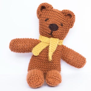 May include: A brown knitted teddy bear wearing a yellow knitted scarf. The bear has black embroidered eyes and a black nose.
