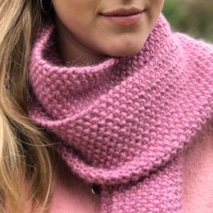 May include: A close-up of a pink knitted scarf worn around the neck of a person. The scarf is made of a soft, fuzzy yarn and has a simple, textured stitch pattern.