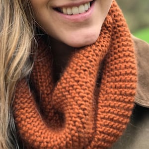 May include: A close-up of a knitted cowl in a warm orange color. The cowl is made of a soft, chunky yarn and has a simple, ribbed texture.