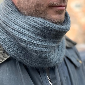 May include: A close-up of a person wearing a blue knitted scarf. The scarf is thick and warm, and it is folded over twice.