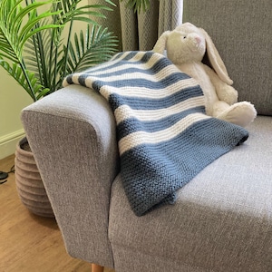 Op de afbeelding: Een blauw-wit gestreepte gebreide deken over de arm van een grijze bank. Een wit pluchen konijn zit naast de deken. Op de achtergrond is een plant in een pot te zien.