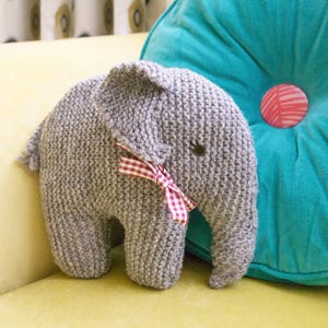 May include: A gray knitted elephant plush toy with a red and white gingham bow. The elephant is sitting on a yellow couch.