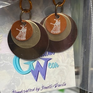 May include: A pair of earrings with a layered design. The earrings are made of three circular metal pieces in shades of brown, silver, and black. Each earring features a small copper disc with a white outline of a Dalek, a fictional robot from the British science fiction television series Doctor Who.