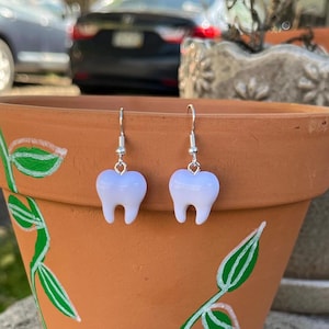 May include: A pair of novelty earrings featuring a white tooth design. The earrings are suspended from silver-colored hooks. The earrings are displayed in front of a terracotta-colored flower pot with green and white painted floral accents.