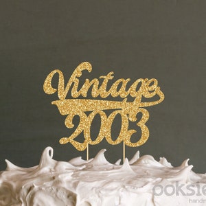 Könnte beinhalten: Ein goldener Glitzer-Kuchenaufsatz mit den Worten "Vintage 2003" in einer Schreibschrift. Der Aufsatz befindet sich auf einem weiß glasierten Kuchen. Der Hintergrund ist einfarbig dunkelgrau.