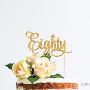 Puede incluir: Topper de pastel de purpurina dorada con la palabra "Eighty" en una fuente cursiva. El topper está encima de un pastel blanco con flores.