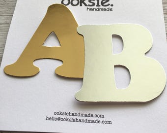 May include: Gold and white letter cutouts of the letters A and B. The letters are on a white card with the text "ooksie. handmade" and the website address "ooksiehandmade.com" and "hello@ooksiehandmade.com".