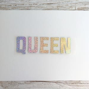 May include: A white card with the word "QUEEN" spelled out in iridescent glitter letters.