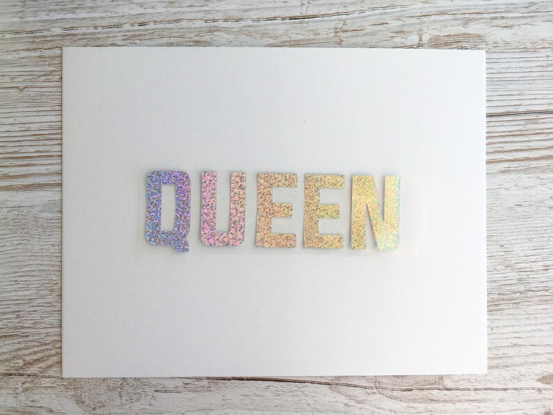 May include: A white card with the word "QUEEN" spelled out in iridescent glitter letters.