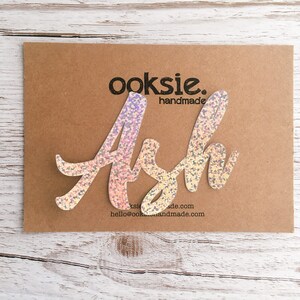May include: A sparkly iridescent pink and white name cutout that says "Ash" with the text "ooksie. handmade" and a website address below.