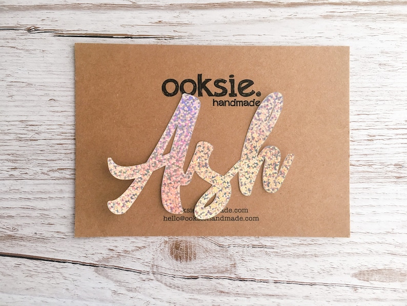 May include: A sparkly iridescent pink and white name cutout that says "Ash" with the text "ooksie. handmade" and a website address below.