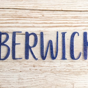May include: Blue glitter vinyl lettering spelling out the word "BERWICK" on a light wood background.